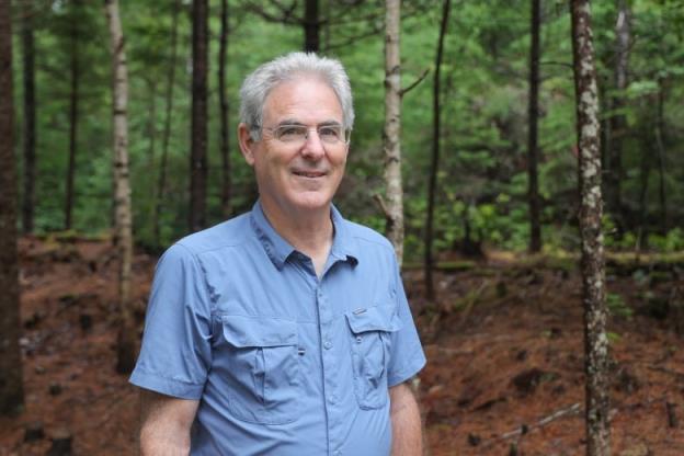 A portrait of a man in a blue collared shirt, wearing glasses and smiling as he looks into the camera with a wooded area as a backdrop.