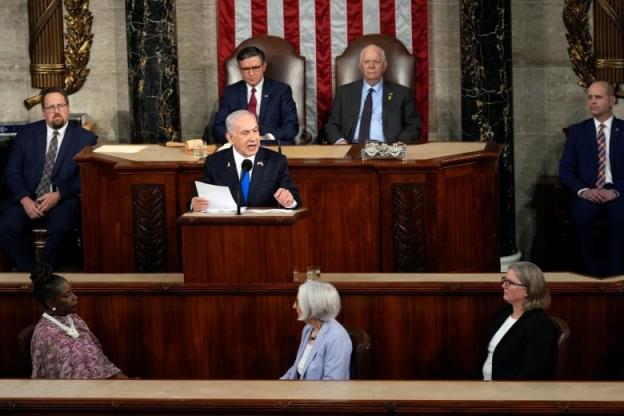 A man in a dark suit speaks while seated at a lectern in a large room decorated with an American flag as others listen. 