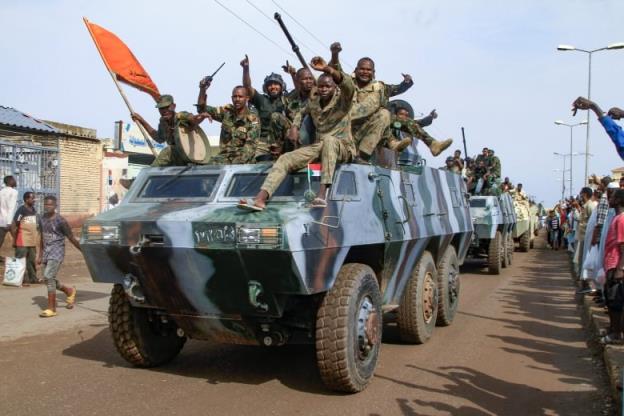 Several men in military fatigues raise their arms and salute atop a tank being driven down a road in what appears to be a parade. o<em></em>nlookers watch or cheer on either side of the road.