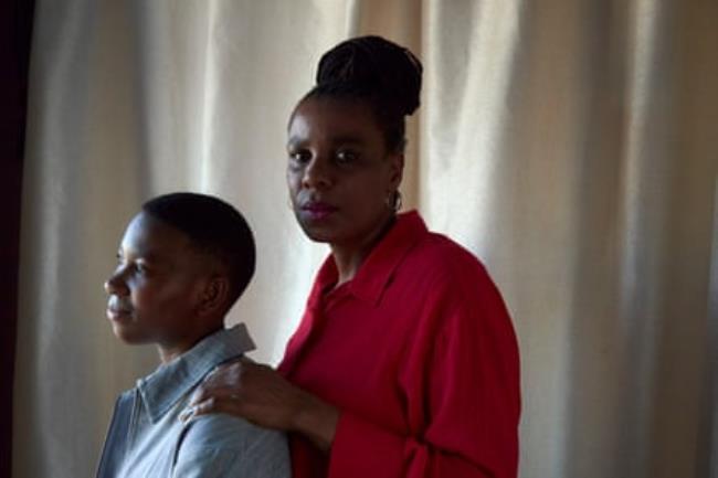 A middle-aged African woman looks at the camera while a teenager sits in front of her and looks away