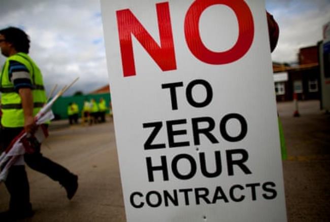 An individual in a hi-vis jacket walking past a sign saying 'No to zero hour contracts' 