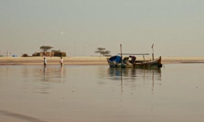 A still from a film shows a boat on calm sea next to a beach. Two people are on the boat, another two wade through the water