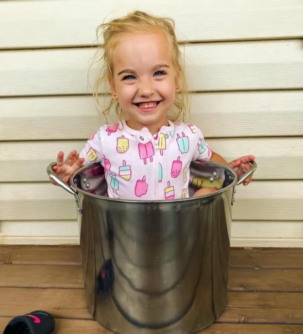 Little smiling blond girl in pink outfit sitting in a large me<em></em>tal lobster pot, smiling