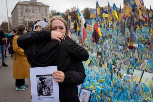 Two women hug as they remember their loved one killed in the war with Russia. One of the women is holding a photo of her dead partner. here are flags and banners and photos of other soldiers laid out behind them.
