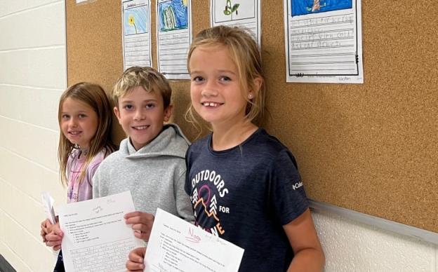 Three smiling elementary students stand in a row in a school hallway, each showing off a written assignment a<em></em>bout local plants. 