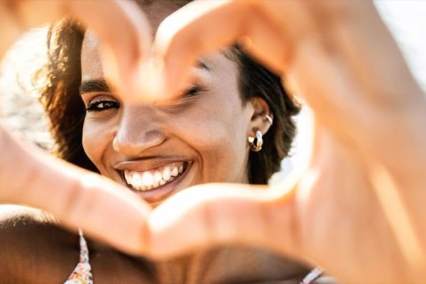 Close up image of smiling woman in swimwear on the beach making a heart shape with hands
