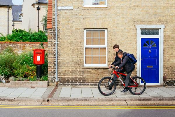 View from the street of two teen boys walking and cycling from school in front of a house