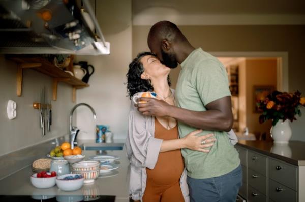 Affectio<em></em>nate pregnant couple kissing while standing in kitchen at home
