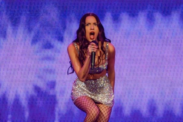 Olivia Rodrigo singing o<em></em>nstage into a microphone wearing a silver glittery skirt and cropped top