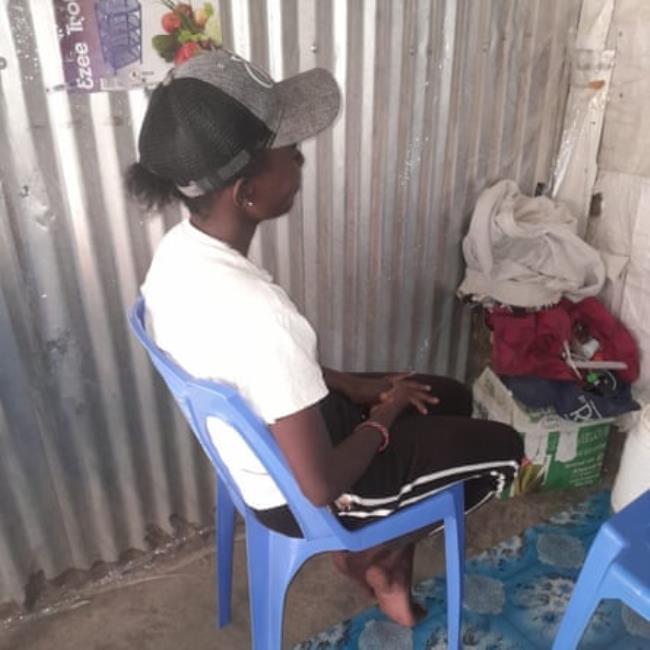 A young woman wearing a ba<em></em>seball cap, white shirt and black trousers with two white stripes sits in a plastic chair next to a corrugated tin wall.