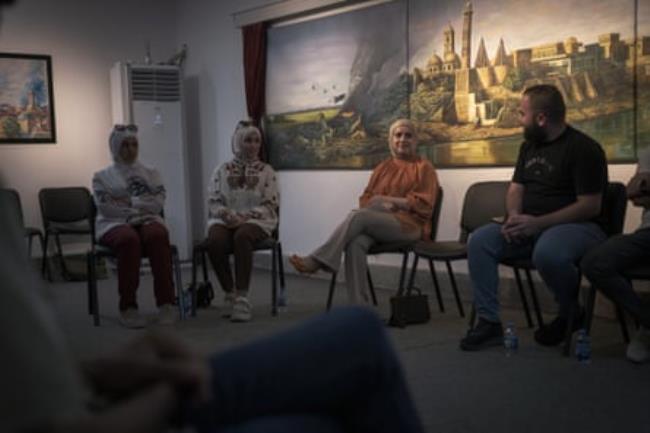 A group of men and women sitting in a circle with a painting of a city in the background