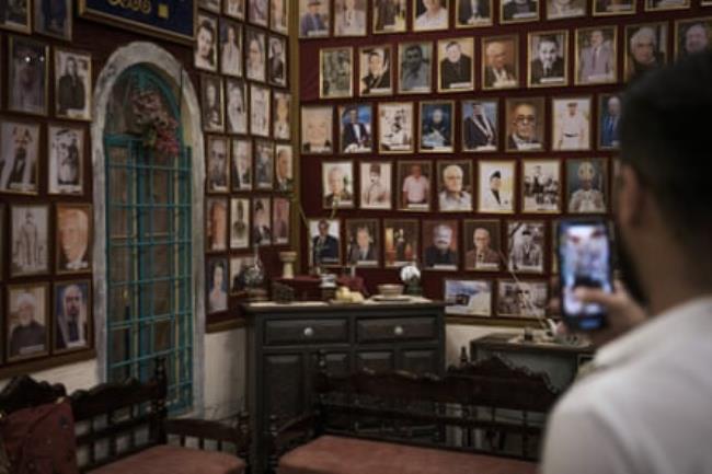 A man takes a photo on his phone of the walls of a room covered with old pictures of mostly men and some women
