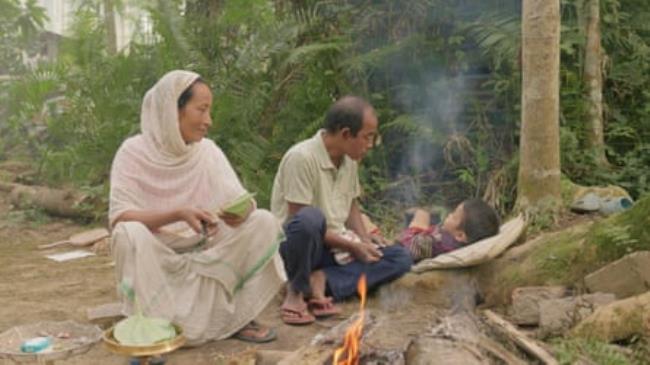 A man, woman and child sit on the ground near a fire next to trees