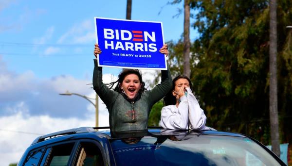 Young supporters cheer from their car holding a Biden-Harris sign.
