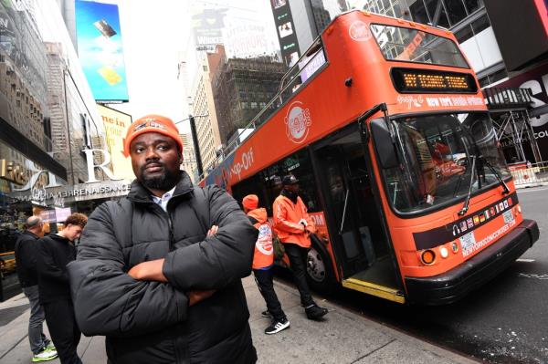 Bamise Jegede, owner of New York Ico<em></em>nic Cruises bus tours, standing in front of a double-decker bus, expressing co<em></em>ncern over intimidation and vandalism from rival company, Top View.