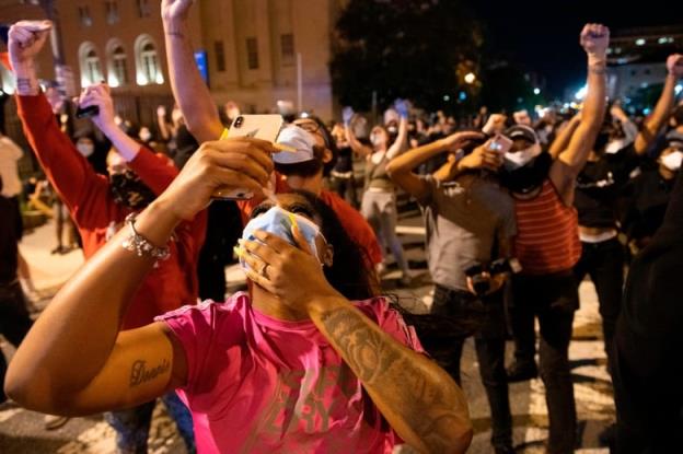 Several people are shown standing outside on a street in a nighttime photo, looking up at the sky. They are wearing COVID-19 masks and some are holding pho<em></em>nes up.