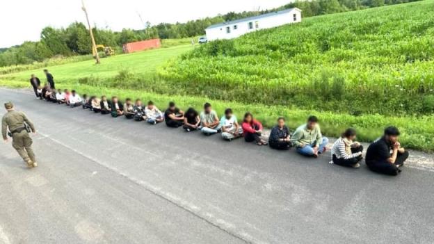 A group of people sit cross-legged on a road as a U.S. Border Patrol agent walks by them.