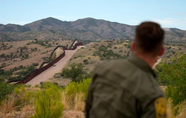 A man with his back to the camera looks across an arid, hilly landscape — marked by a long, high wall. 