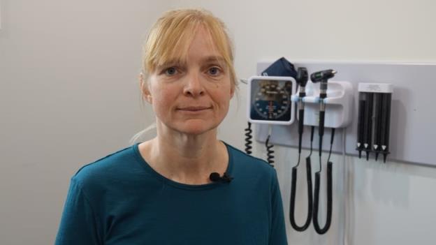 A woman with blo<em></em>nde hair and a blue shirt stands in a white room with medical equipment, including a blood pressure gauge and otoscopes from examining a patient's ears, hanging on the wall behind her.
