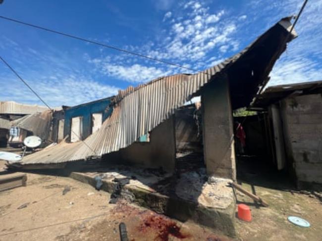 A corrugated iron roof collapses over a co<em></em>ncrete wall with bloodstains on the dirt in front of it.