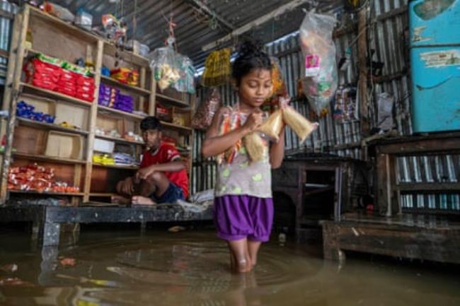 A child carrying food walks knee-deep in water in a flooded rural shop.