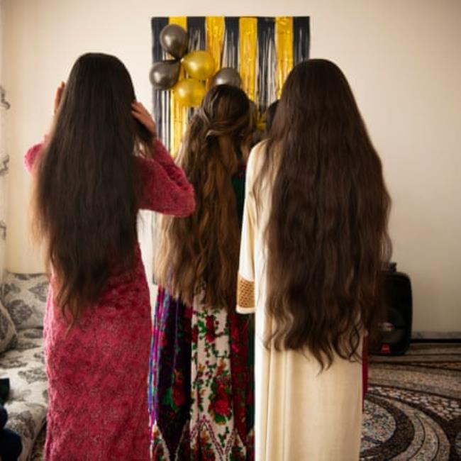 Three unveiled women with long hair wearing colourful dresses stand in a room with their backs to us. Balloons and streamers can be seen in the background