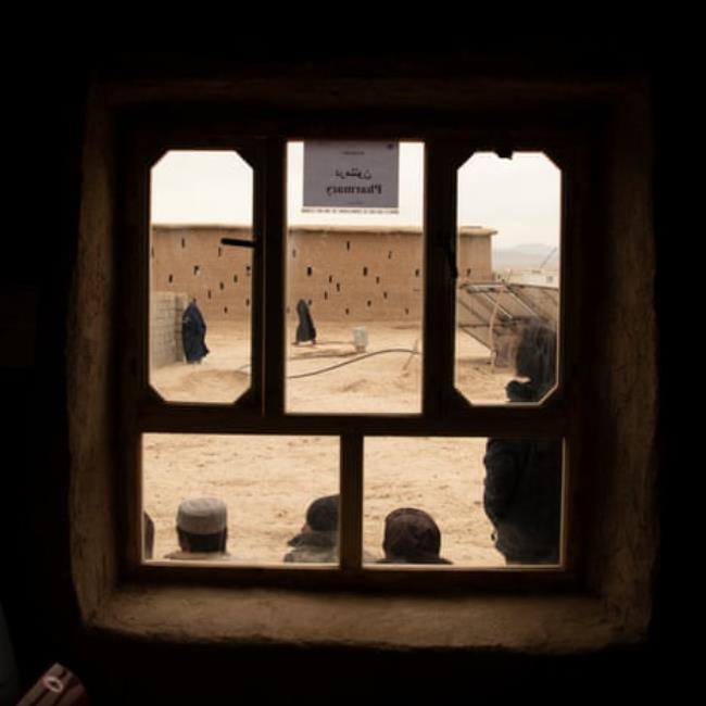 A view through a window into a dusty courtyard with men sitting at one end and two women in burqas standing at the other. A sign on the window reads ‘pharmacy’