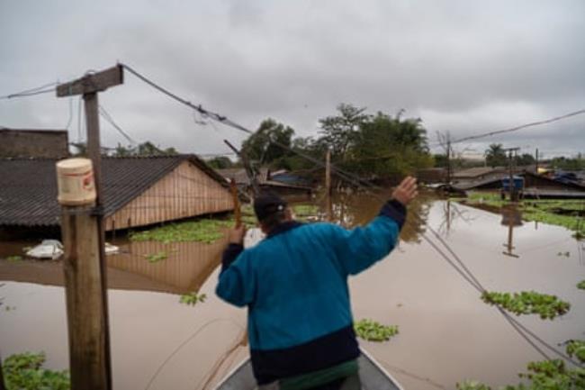 A man seen from behind as he uses a pole to steer his small boat towards buildings wher<em></em>e water is almost up to the eaves 