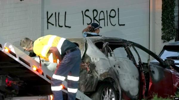 FILE - This f<em></em>rame grab taken from video footage provided by the Australian Broadcast Corporation on December 11, 2024 shows a police officer walking past a torched car being removed in front of anti-Israel graffiti written on a wall in the Sydney suburb of Woollahra.