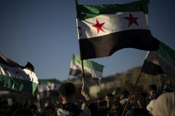 Syrian flags fly against a blue sky, held in the hands of people marching together