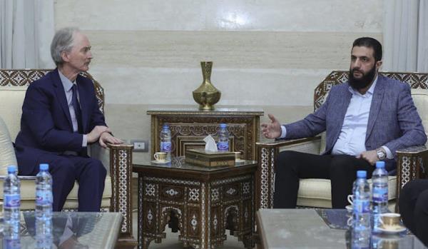 A white man in a suit sits across from a bearded Middle Eastern man in a blazer and dress shirt
