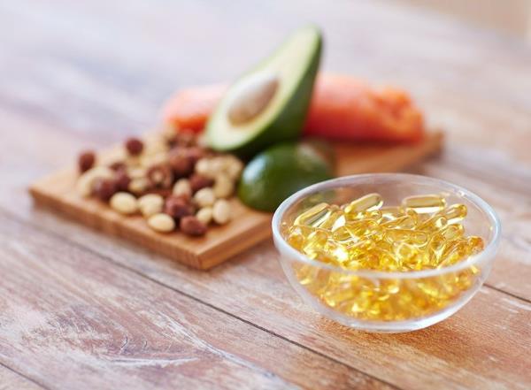 omega 3 fish oil supplements in a clear bowl next to a display of nuts, avocado, and salmon, all on a wood background