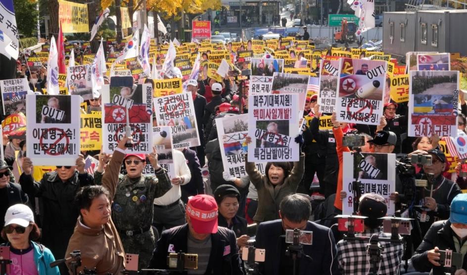 A crowd of people are outside, with trees with yellow leaves in the distance at the side of the road they seem to be standing on. They are holding signs above their heads. 