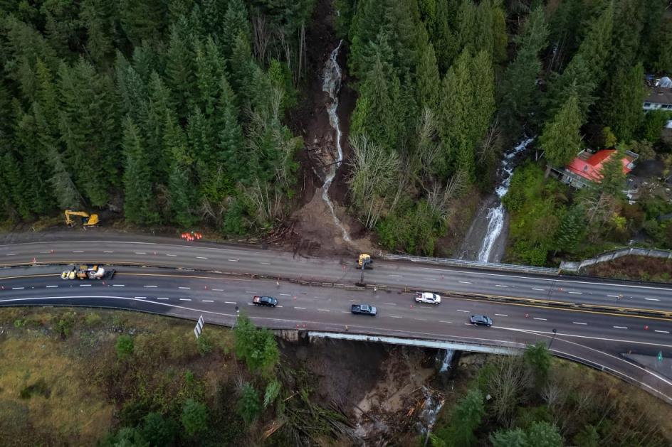 An aerial view of a highway that has had mud and debris over on one side of it, with a landslide clearly visible along a slope.