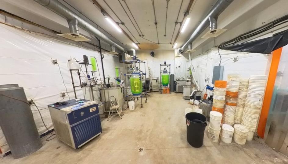 The interior of what looks like a lab with various co<em></em>ntainers and machines on a wooden floor under large hood vents and fluorescent lights.