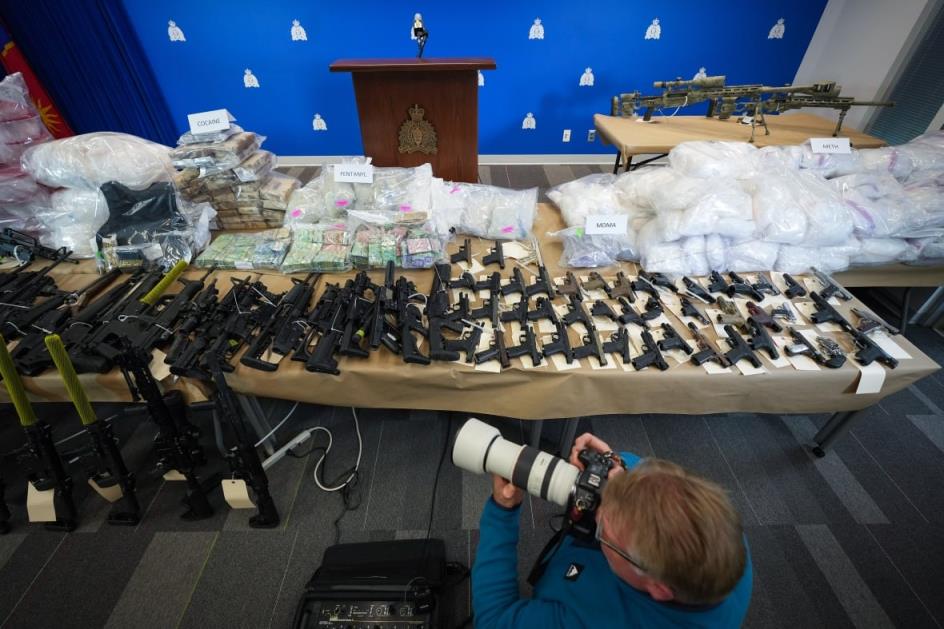 A table full of guns, plastic baggies with powder inside, body armour and other paraphernalia. A photographer is seen in the foreground.