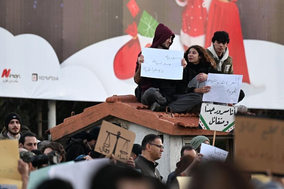 Three young people sit on the roof of a hut in a square holding placards in Arabic.
