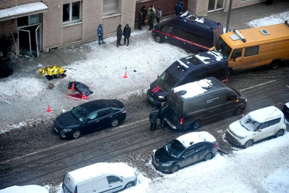 People stand around on a snowy sidewalk outside an apartment building. Tarpaulin cover two objects on the ground.