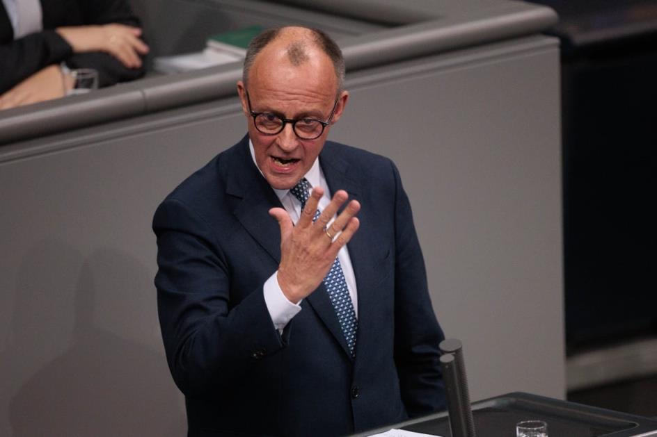 A balding, clean shaven man wearing glasses and a suit and tie speaks while standing in what looks like a government chamber.