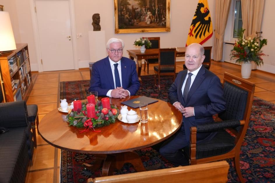Two older men in suits sit at a table inside an ornate room with a flag, portrait and flowers. A bound book rests on a table wher<em></em>e the two men are sitting.