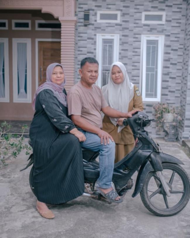 Two women and a man pose on a motorbike in front of a house