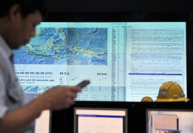 A man checks his phone against a backdrop of a large screen showing meteorological information