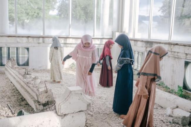 Five girls in head scarves and long dresses play among the rubble of a mosque