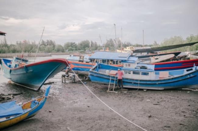 A man sitting next to a fishing boat among a group on boats on the shore