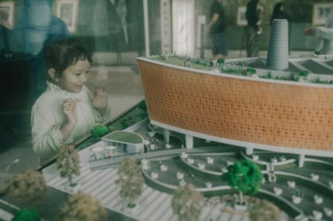 A small child smiles at a model of a building in a glass display case