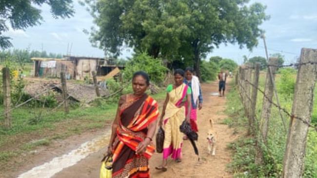 Three women in colourful saris carrying axes and bags walk along a sandy lane past a hut