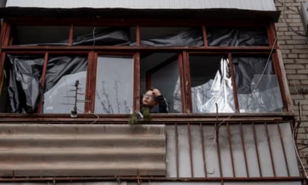 A boy looks out of a damaged window; most of the windows in a block have been blown out with some covered by plastic, and the front of the building has lost some of its iron cladding