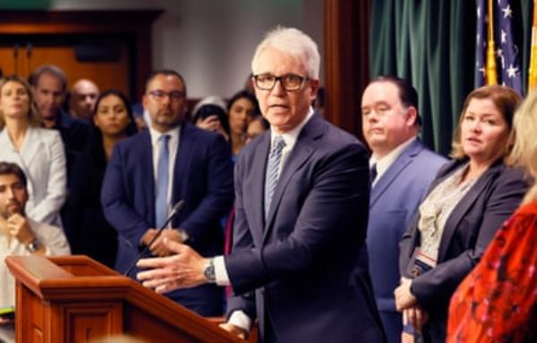 a man wearing a suit speaks at a lectern, a group of people looking on behind him
