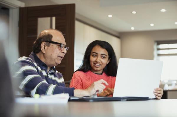 A grandfather is helping his granddaughter as she works from a laptop; they look happy to be bo<em></em>nding and working together.