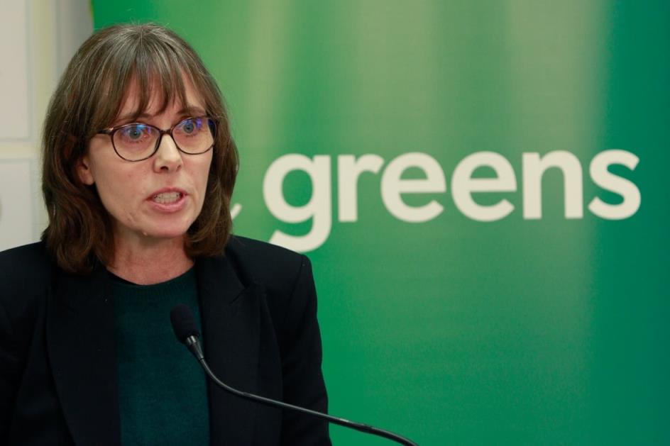 A woman speaks in front of a screen that reads 'Greens'.
